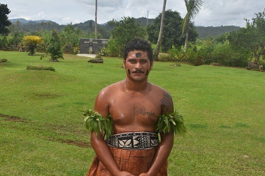 A person in traditional attire with a lei, standing in a grassy area with trees and a building in the background. 3