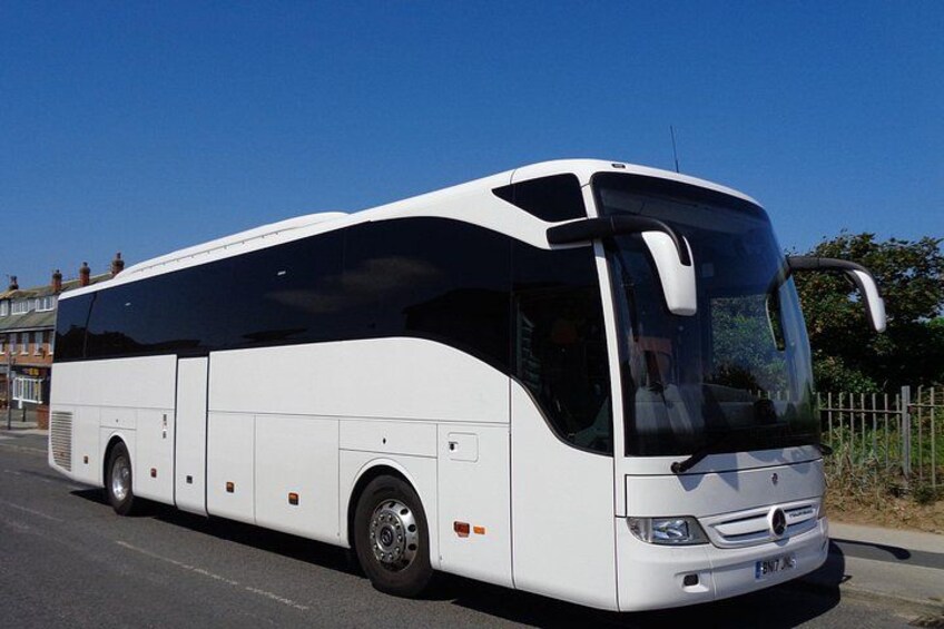 A white bus with a large windshield and side windows, parked on a road with buildings and a fence in the background. 5