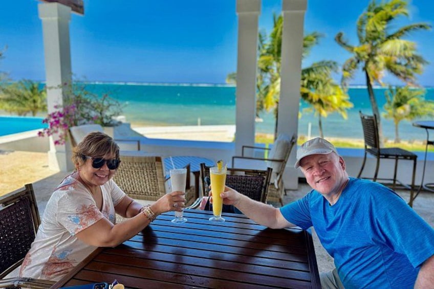 Two people toasting with drinks at a wooden table with a view of the ocean and palm trees. 2