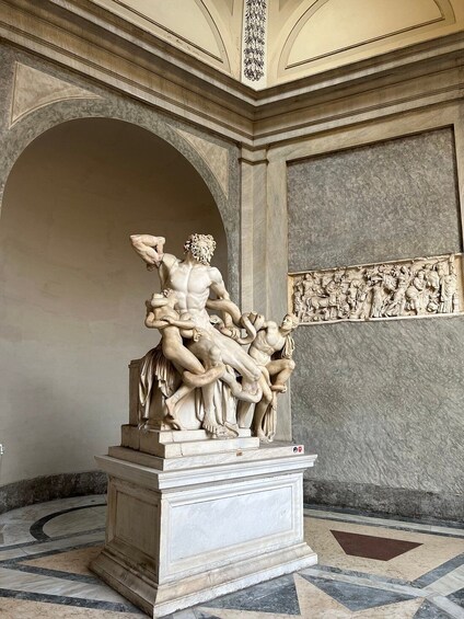 Une sculpture en marbre représentant une scène mythologique, avec des personnages dans des poses dynamiques, le tout se détachant sur un fond composé d’éléments architecturaux classiques. 3