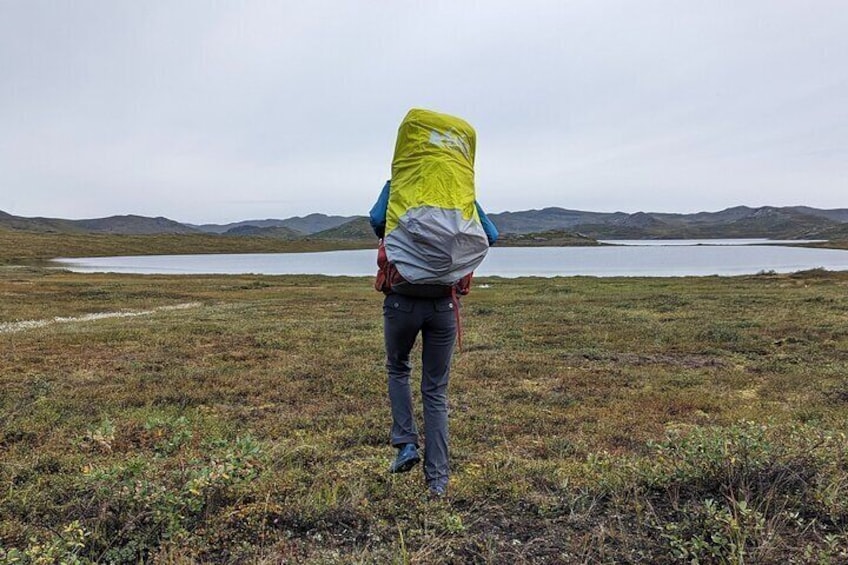 A person walking on a grassy field with a large backpack, a lake, and hills in the background. 5