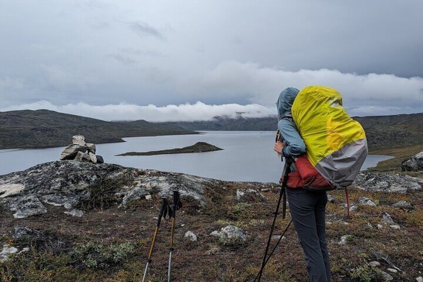 A hiker with a yellow and gray backpack standing on rocky terrain overlooking a lake and distant hills. 1