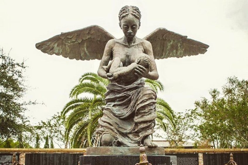Una estatua de un ángel sosteniendo a un niño, rodeada de palmeras y un muro de piedra. 3