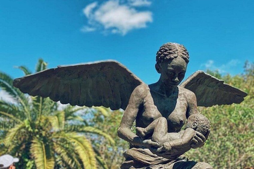 Une statue d’un ange aux ailes déployées, tenant un enfant dans ses bras, se dressant sur fond de palmiers et d’un ciel dégagé. 3