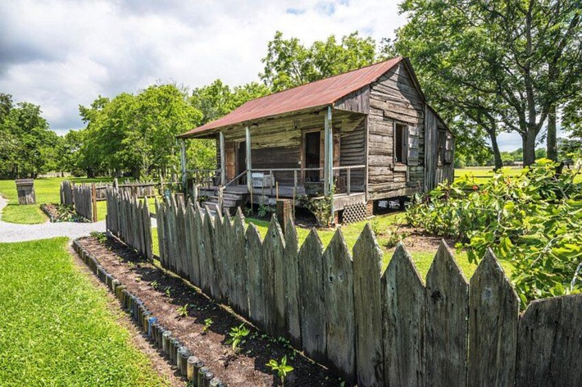 A rustic wooden cabin with a porch, surrounded by a wooden picket fence and lush greenery. 1