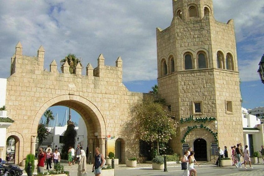 Une porte historique en pierre, surmontée d’une tour, entourée de piétons et de verdure. 2