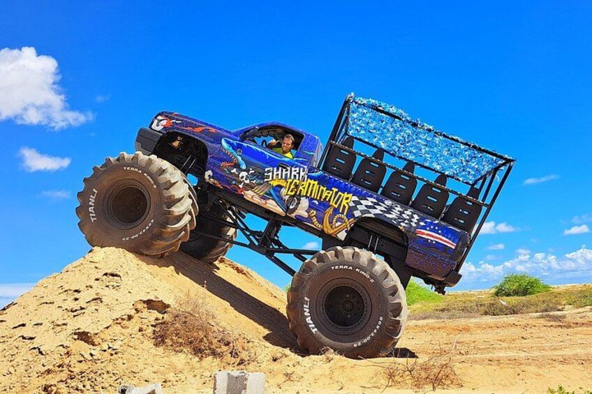 A blue monster truck with a raised bed, driving on a sandy terrain. 1