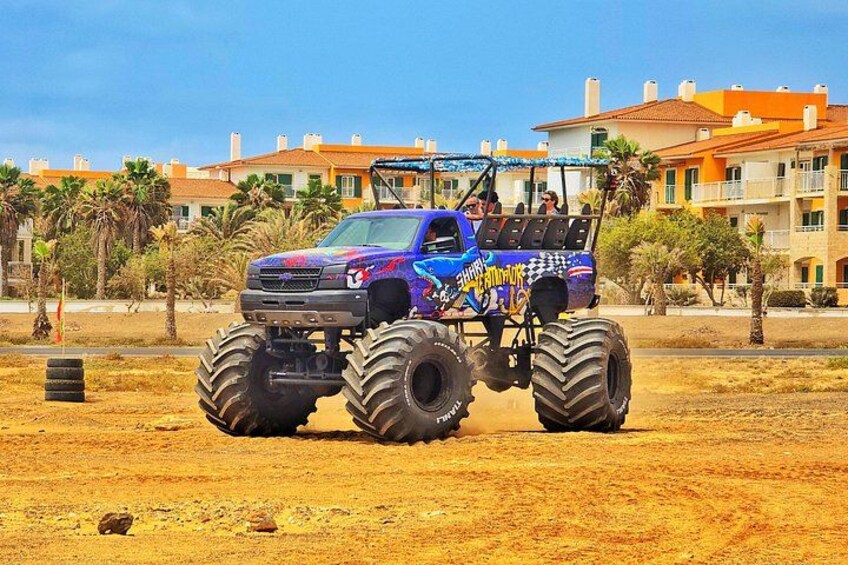 A large, colorful monster truck with oversized tires parked in a desert-like area with palm trees and buildings in the background. 3