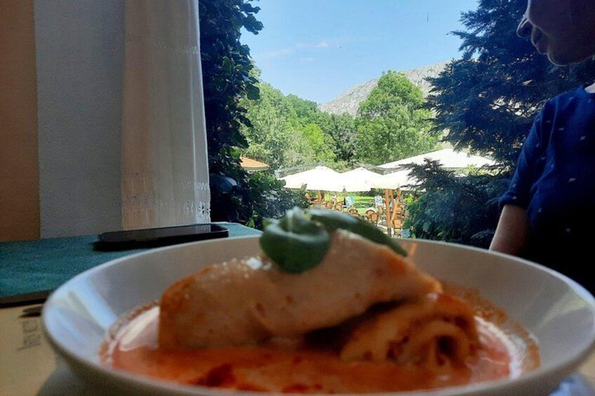 A bowl of food with a green leaf garnish, a white plate, and a view of a green landscape with trees and a building in the background. 2