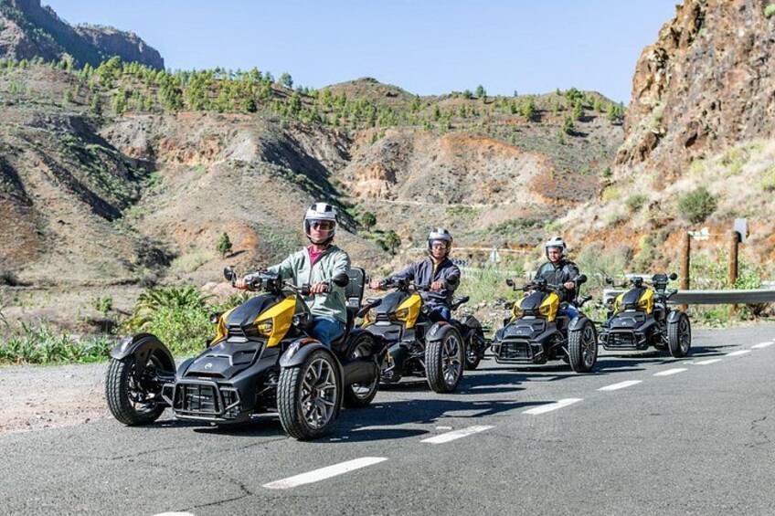 Four people riding quad bikes on a road with a mountainous landscape in the background. 1