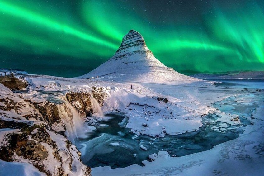 Ett snötäckt landskap med ett vattenfall och ett berg under norrskenet. 1