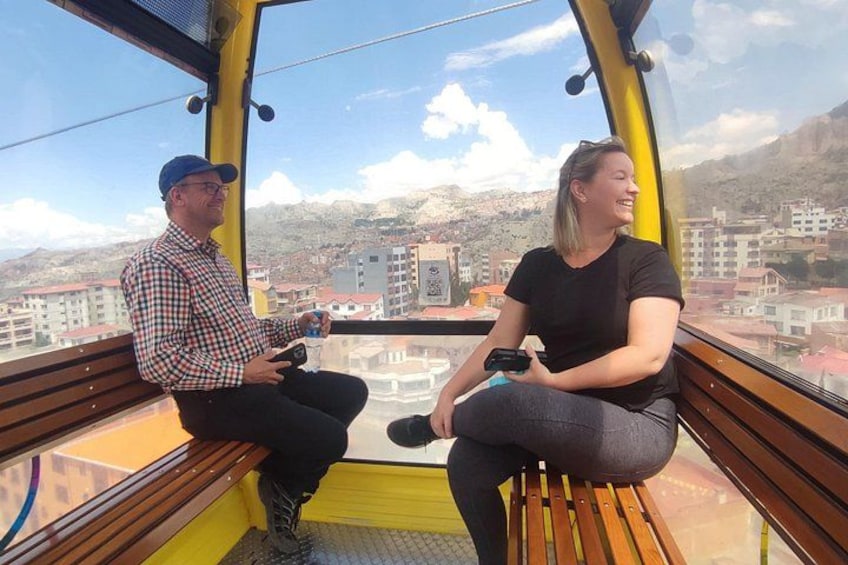 Two people sitting on a bench inside a yellow structure with a view of a cityscape and mountains in the background. 3