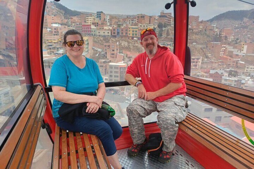 Two people sitting on a bench inside a cable car with a cityscape in the background. 3