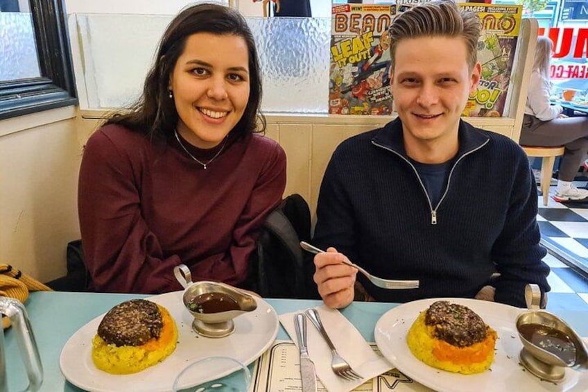 To personer sidder ved et bord med tallerkener mad, et vindue og en plakat i baggrunden. 2