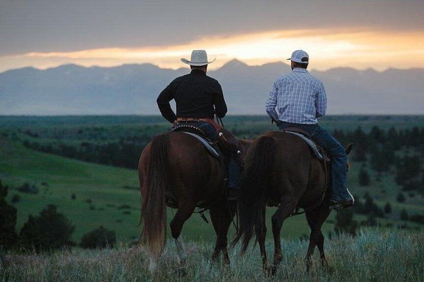Two people riding horses on a grassy hill with a mountain range in the background. 3