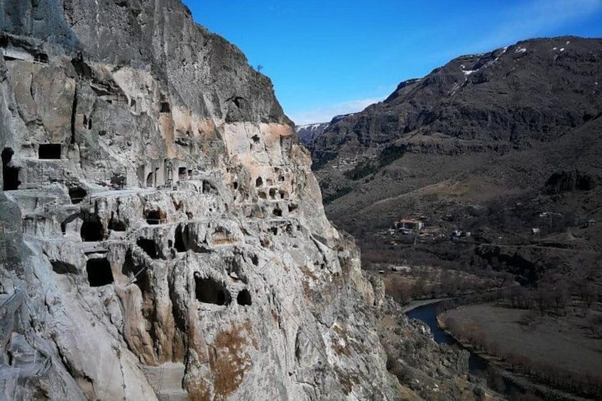 A rocky cliff with multiple cave entrances, a valley, and distant buildings. 1