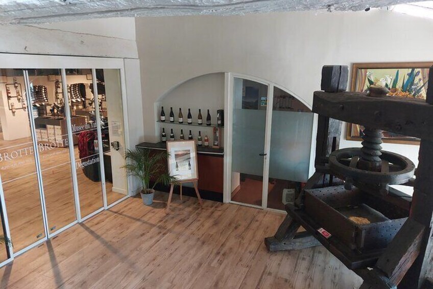 A wine shop interior with wooden floors, a display of wine bottles, and a large mechanical press. 3