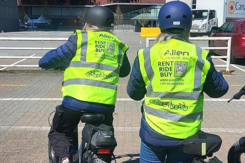 Two individuals on bicycles wearing reflective vests with 'Alien Rent Ride Buy' branding. 4