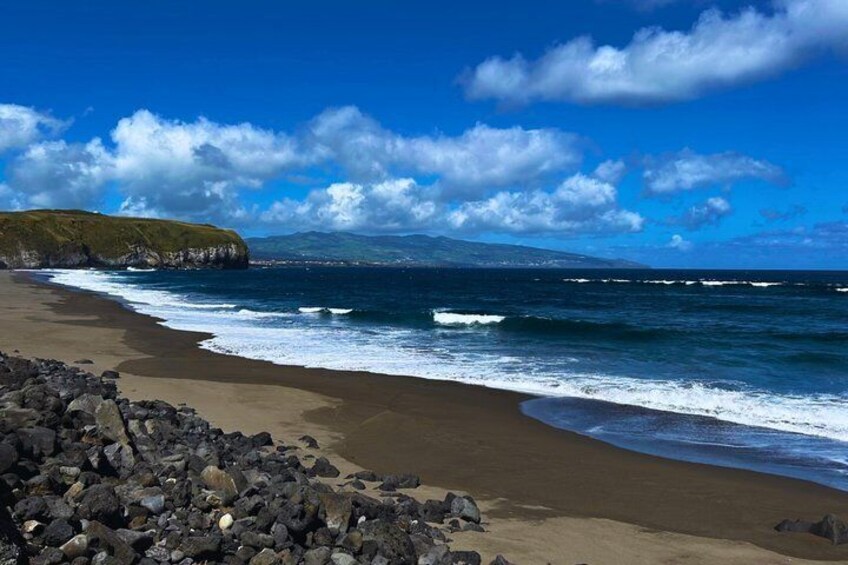 Une plage avec des vagues, un premier plan rocheux et une chaîne de montagnes lointaine sous un ciel bleu. 2