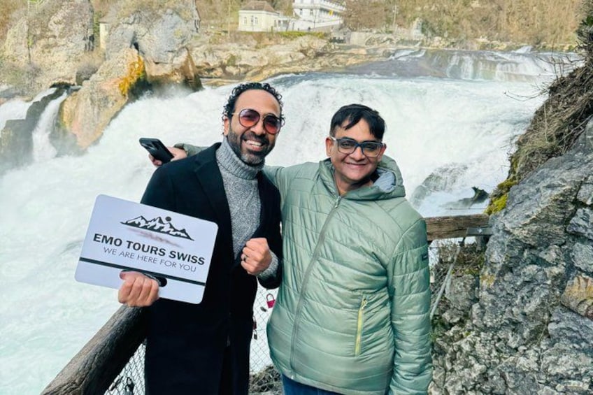 Dos personas de pie junto a una cascada con un letrero que dice 'EMO TOURS SWISS'. 1