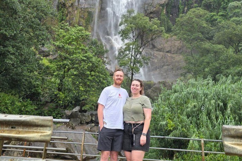 Due persone in piedi di fronte a una cascata, con vegetazione lussureggiante sullo sfondo. 2