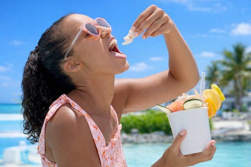 A person holding a drink with a straw and a slice of orange, wearing a sleeveless top, with a beach and palm trees in the background. 3