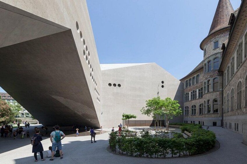A plaza with a large concrete structure, a traditional building with a tower, and a circular garden with trees. 2