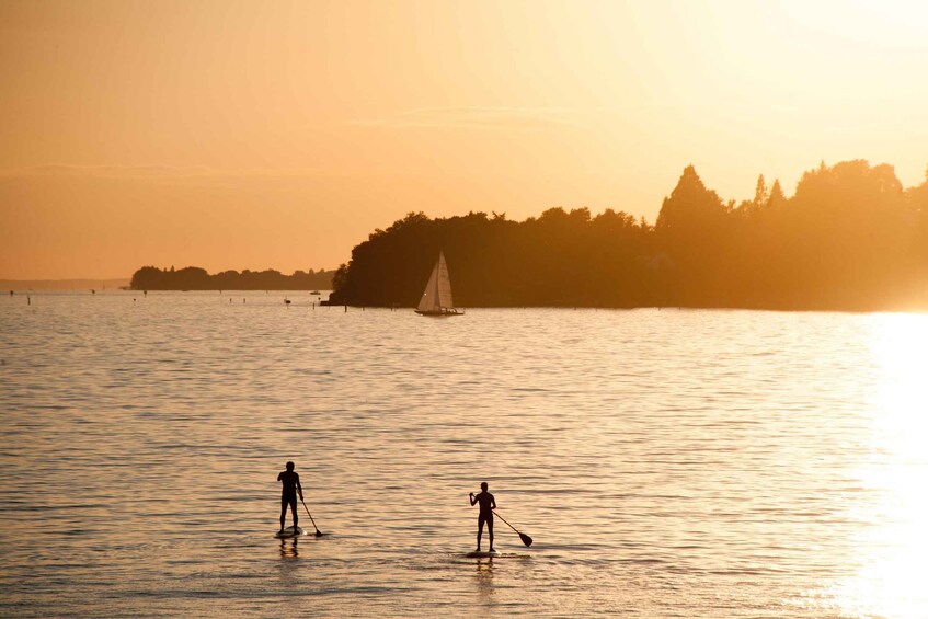 Two people paddleboarding on calm water with a sailboat in the distance and a forested island on the horizon. 2