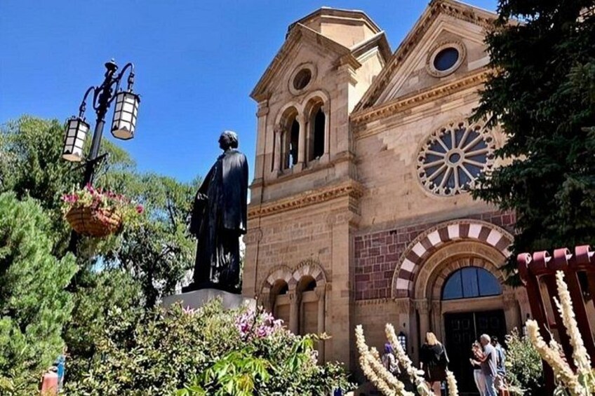 En historisk bygning med et markant clock tower, en statue foran og frodigt grønt område. 1