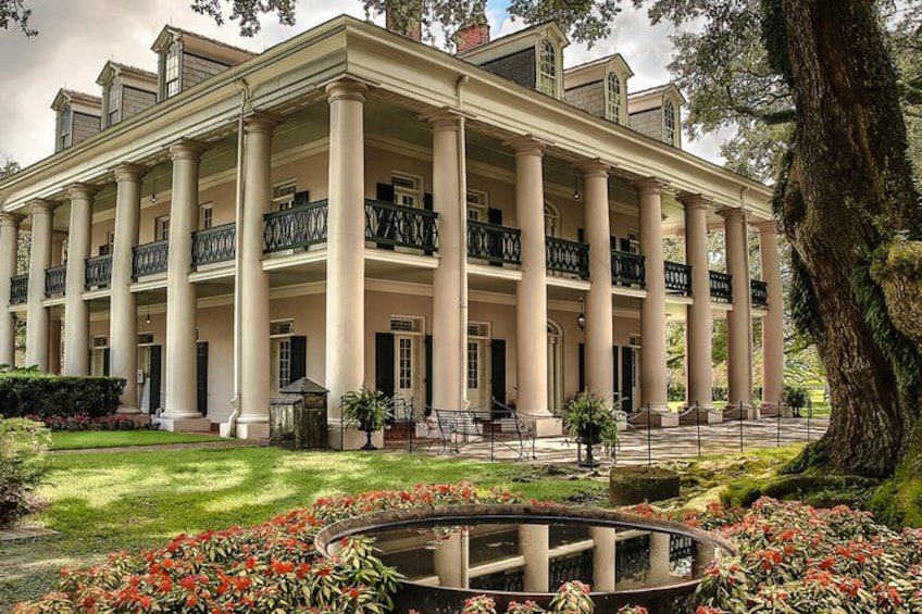 Un ampio edificio in stile coloniale, con numerose colonne e balconi, circondato da un giardino curato e uno stagno. 1