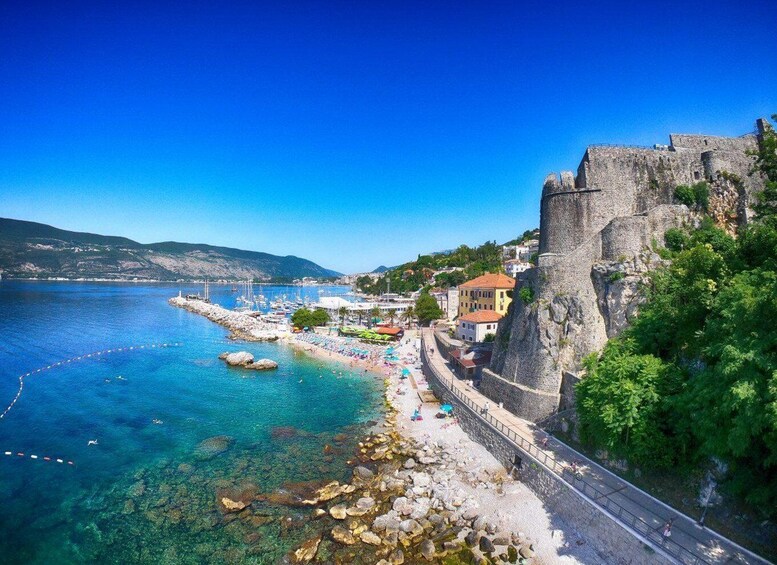 A coastal town with a historic stone wall, clear blue sea, rocky shoreline, and colorful beach umbrellas. 5
