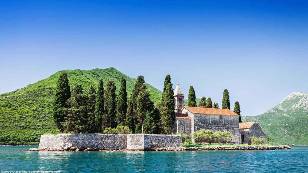 A stone church with a bell tower by a calm sea, surrounded by lush green hills. 3
