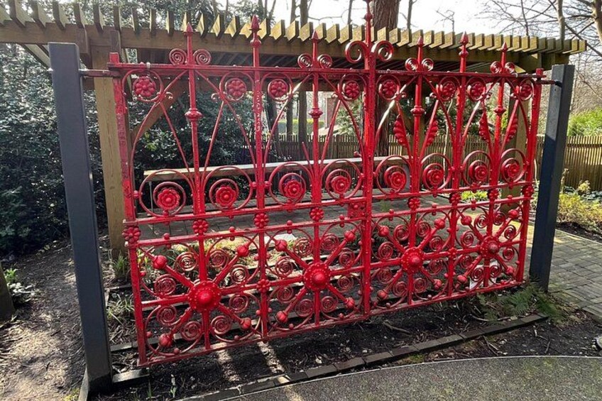 A red wrought iron gate with intricate designs, set in a garden with a wooden pergola overhead. 4
