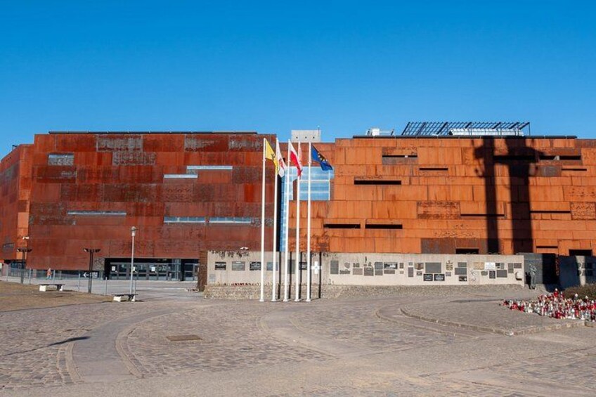 Een groot, rood gebouw met een modern ontwerp, een wimpelstok met verschillende vlaggen en een verhard gebied ervoor. 5