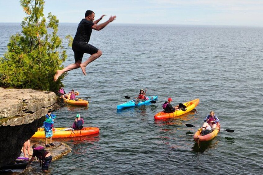 A person jumping into the water from a cliff, kayakers on the water, and a rocky shoreline. 2