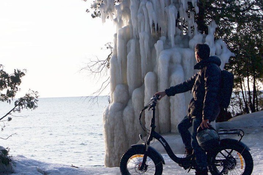 A person on a bicycle with a backpack, near a large ice formation by the sea. 4