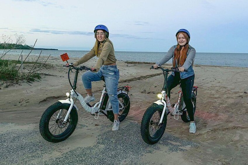 Two people on electric bikes on a sandy beach with the sea in the background. 3