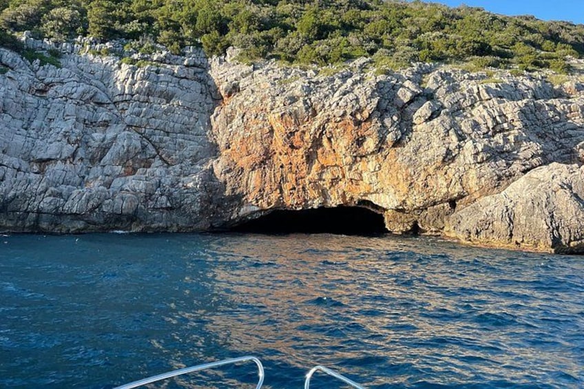 A rocky coastline with a cave entrance, calm sea, and a forested hill in the background. 2