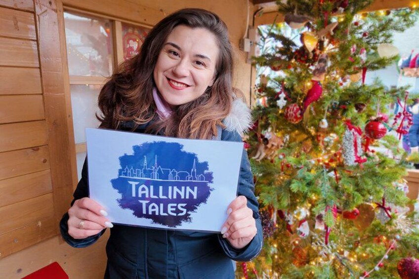 A person holding a sign with 'Tallinn Tales' written on it, standing in front of a decorated Christmas tree. 4