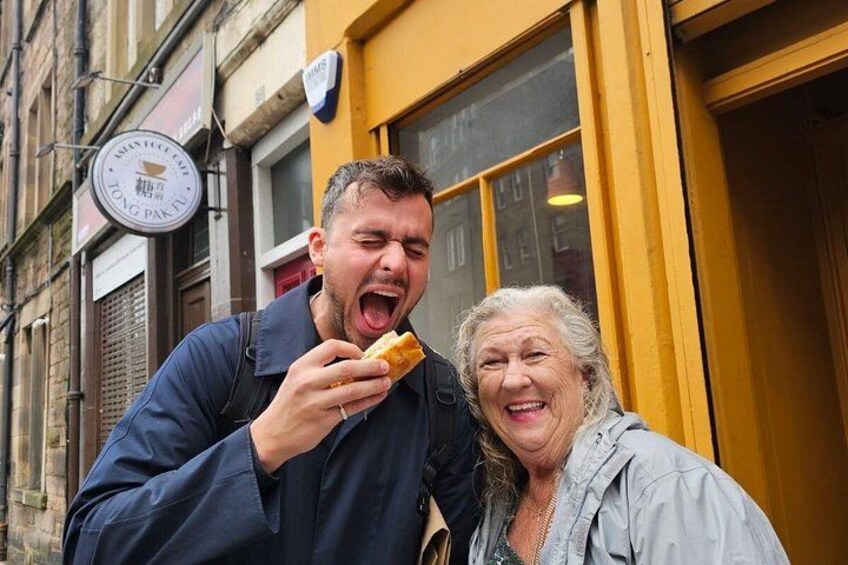 Un homme mange un sandwich et une femme âgée se tient devant un bâtiment sur lequel on peut lire l’inscription « TONG PAK POO ». 3