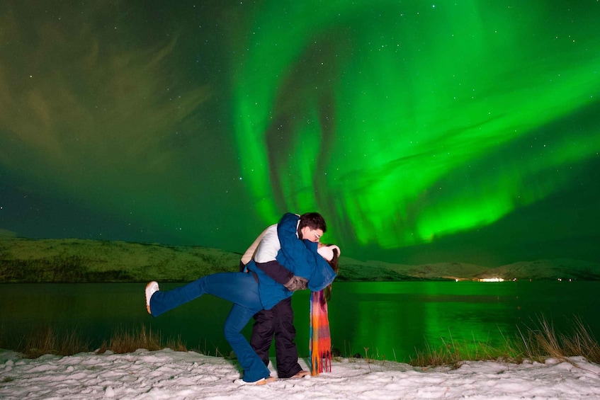 A person in blue and white clothing embracing another person in a colorful scarf, with the Northern Lights in the background. 1