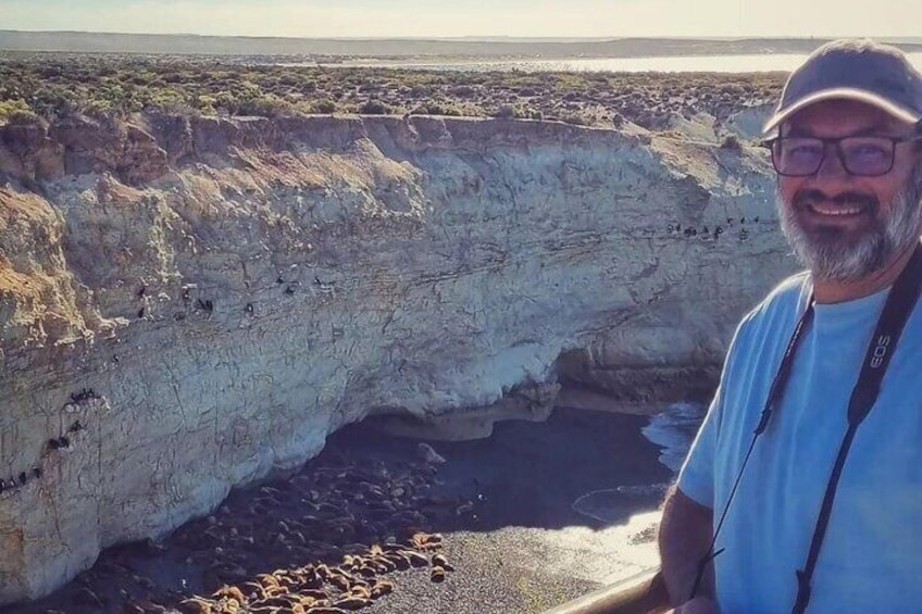 Una persona de pie junto a un acantilado enorme con una pequeña cueva en la base. 1
