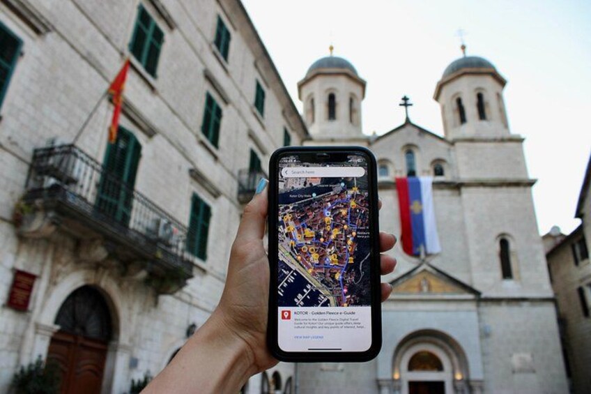 Una mano sosteniendo un teléfono inteligente con una aplicación de mapas, frente a un edificio histórico con torres en forma de cúpula y banderas. 4