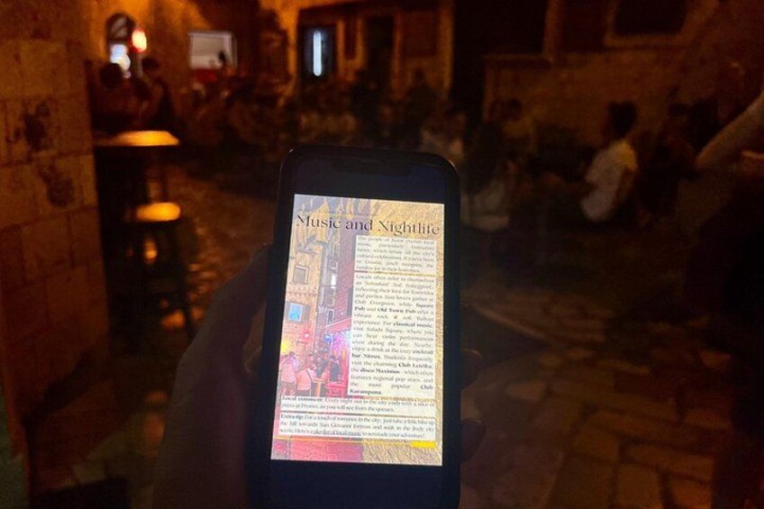Una persona sosteniendo un teléfono inteligente que muestra un artículo titulado 'Música y Vida Nocturna' en un ambiente interior tenuemente iluminado con paredes de ladrillo y gente de fondo. 3