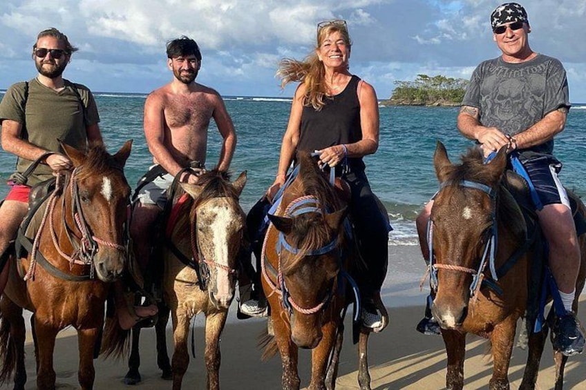 Vier personen paardrijden op een strand, met de oceaan op de achtergrond. 1