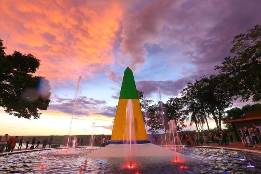 A water fountain with a green, yellow, and blue base, surrounded by trees and a sunset sky. 5