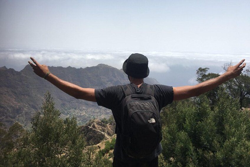 A person standing with arms outstretched on a mountain, facing a valley with trees and a distant mountain range. 5