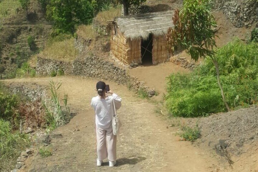 Eine Person fotografiert eine traditionelle, mit Stroh gedeckte Hütte, die von Grünpflanzen gesäumten Feldweg. 4