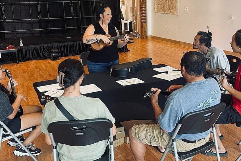 A person playing guitar in a room with a group of seated individuals. 3