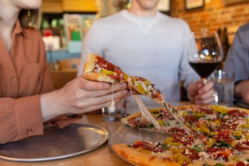 A person holding a slice of pizza with various toppings, a pizza on a tray, and a glass of red wine on the table. 5